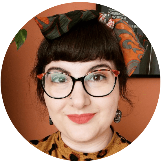 A photograph of the artist Charlotte Thomson-Morley - a white woman with glasses and brown hair tied up with a heady fringe. She is standing against an orange background with a plant.